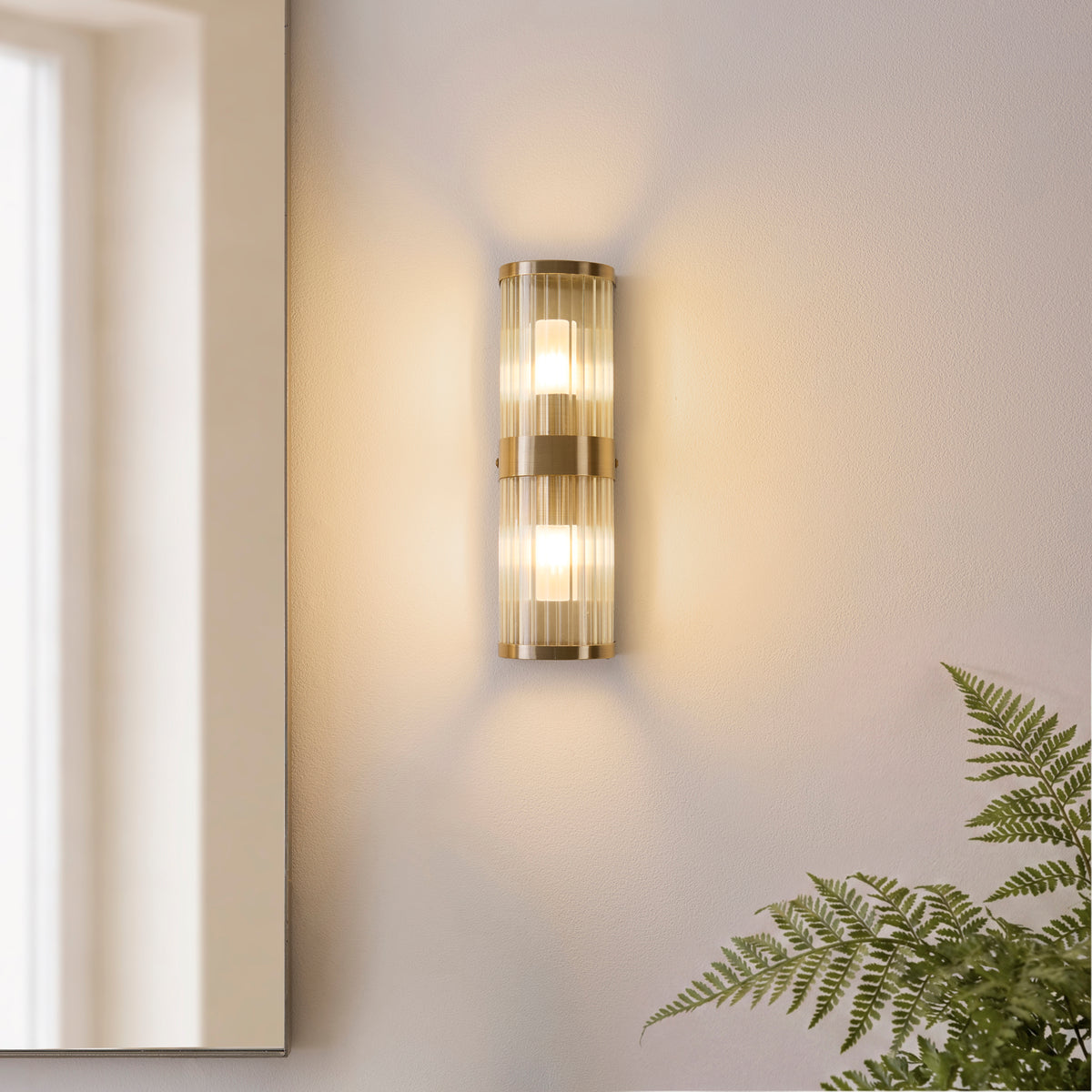 Ridgeton brass wall light casting warm light on a neutral wall beside a fern and mirror