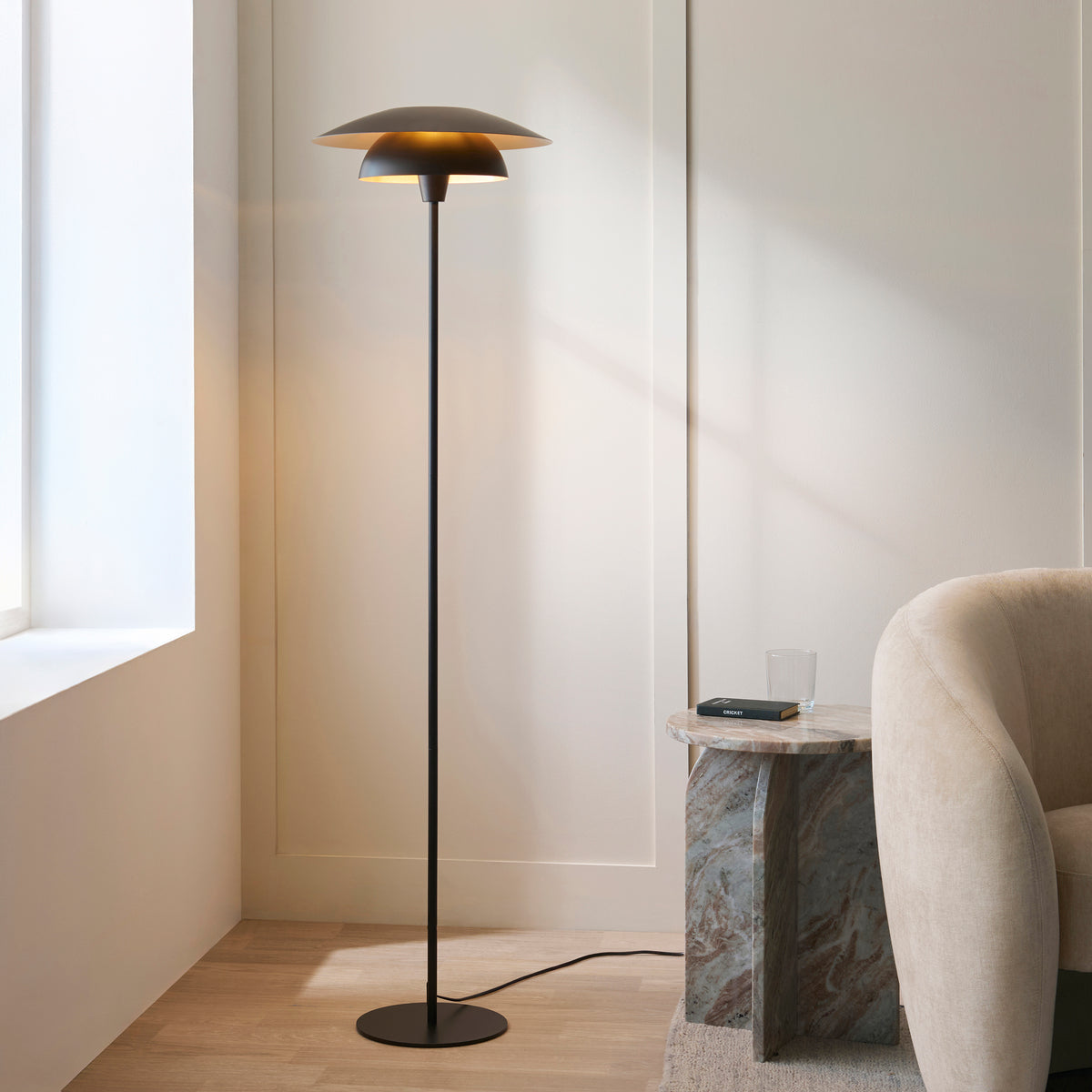 Lyon black floor lamp lit beside a cream armchair and marble side table in a bright living room