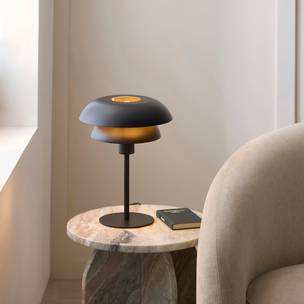 Lyon black table lamp lit on a marble side table with a book beside a cream armchair