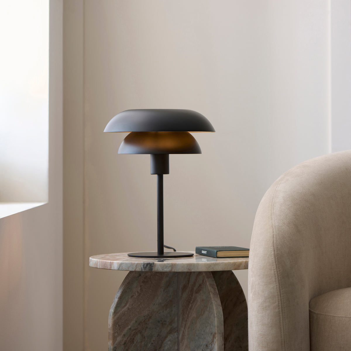 Lyon layered table lamp glowing on a stone side table next to a boucle armchair