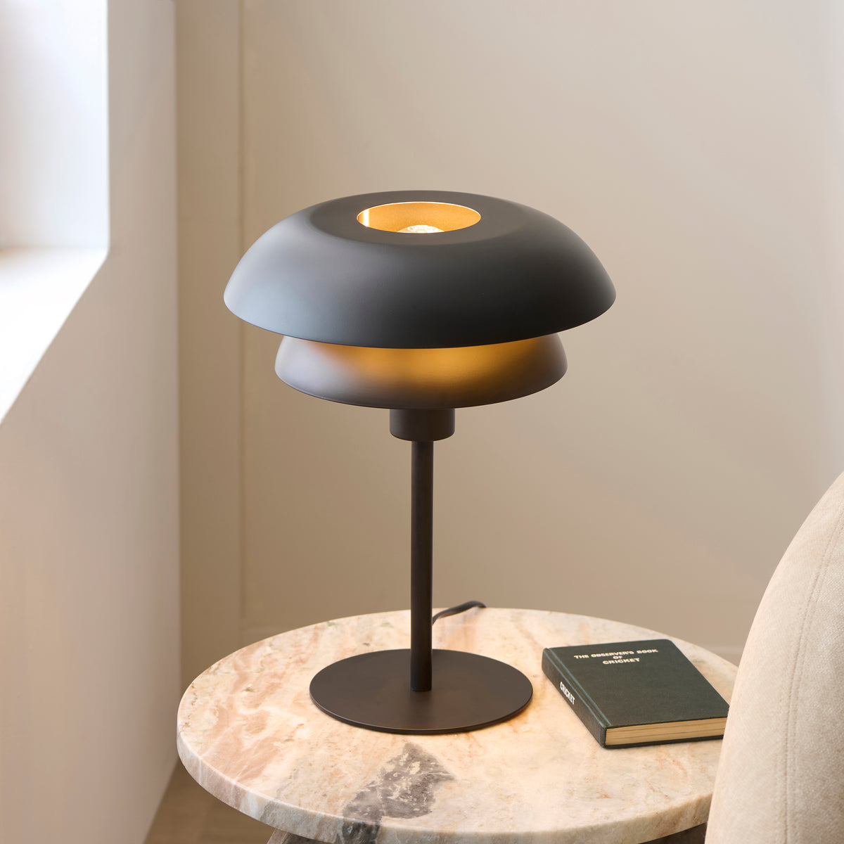 Lyon black and brass table lamp lit on a marble table beside a sunlit window