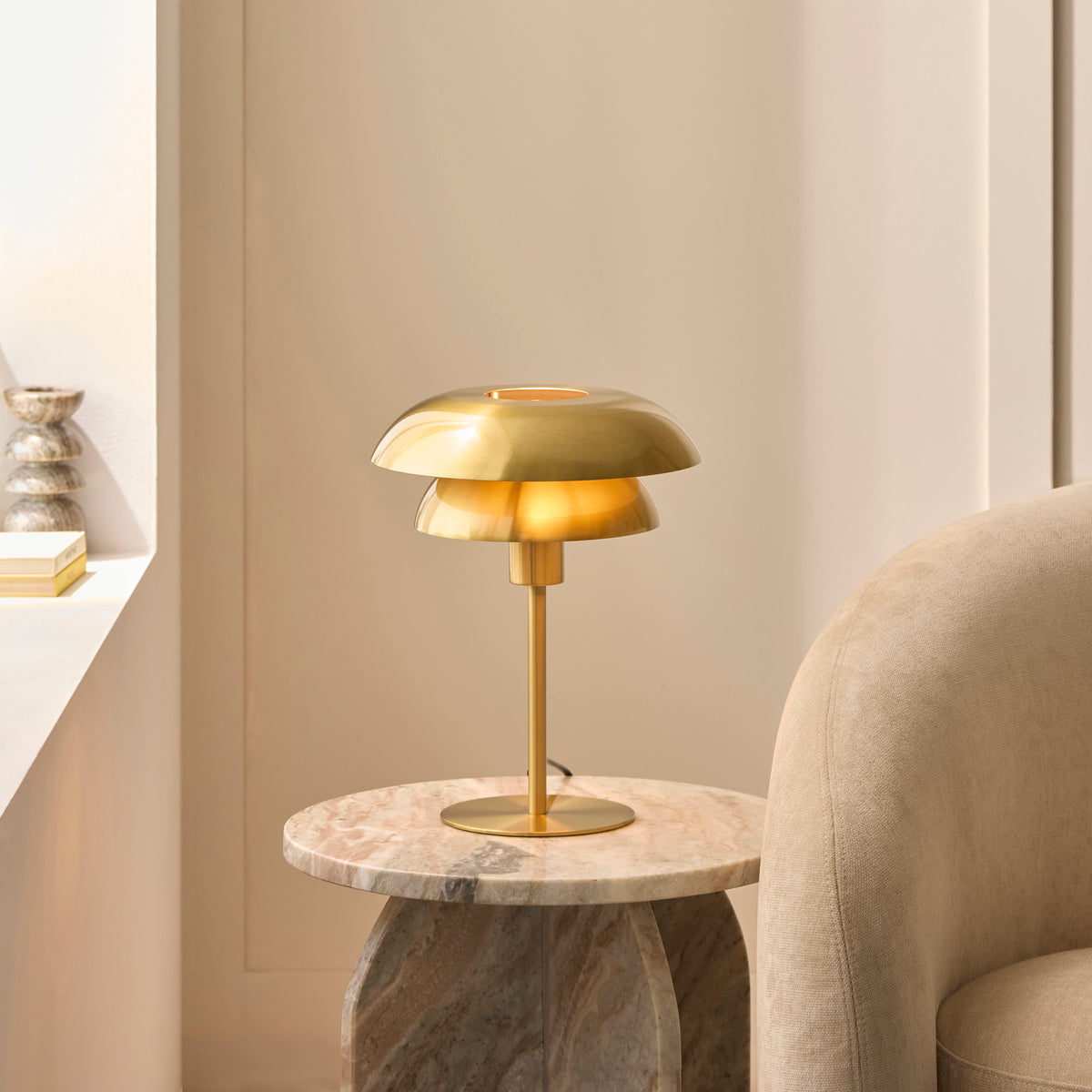 Lyon brass table lamp glowing on a marble side table beside a cream armchair