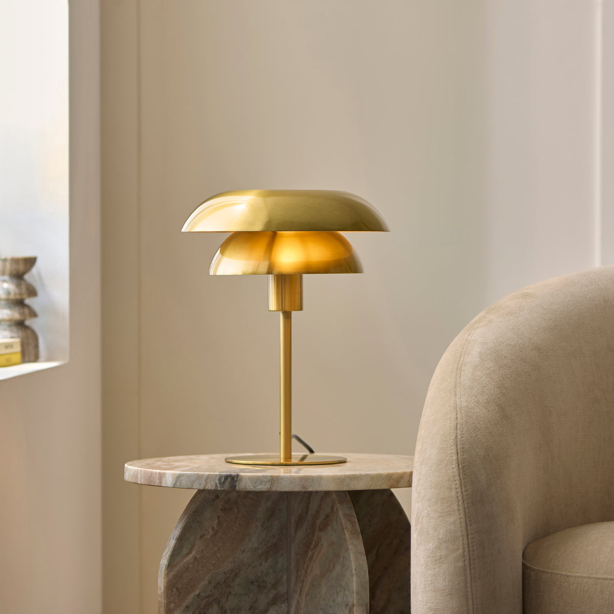 Lyon layered brass table lamp lit on a stone pedestal table next to a boucle chair