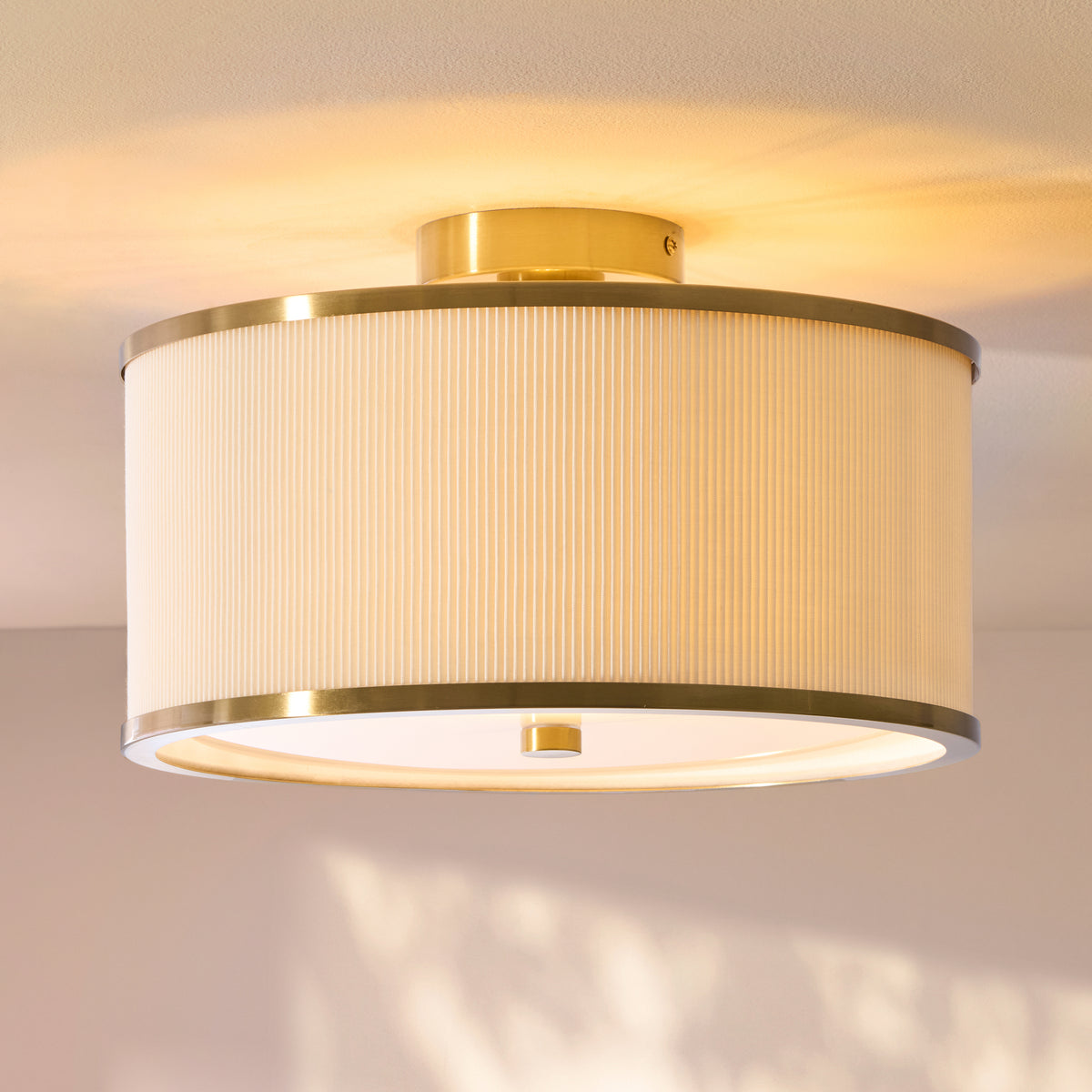 Lexi flush fitting lit close up, showing brass trim, pleated ivory shade and sunlight on the ceiling