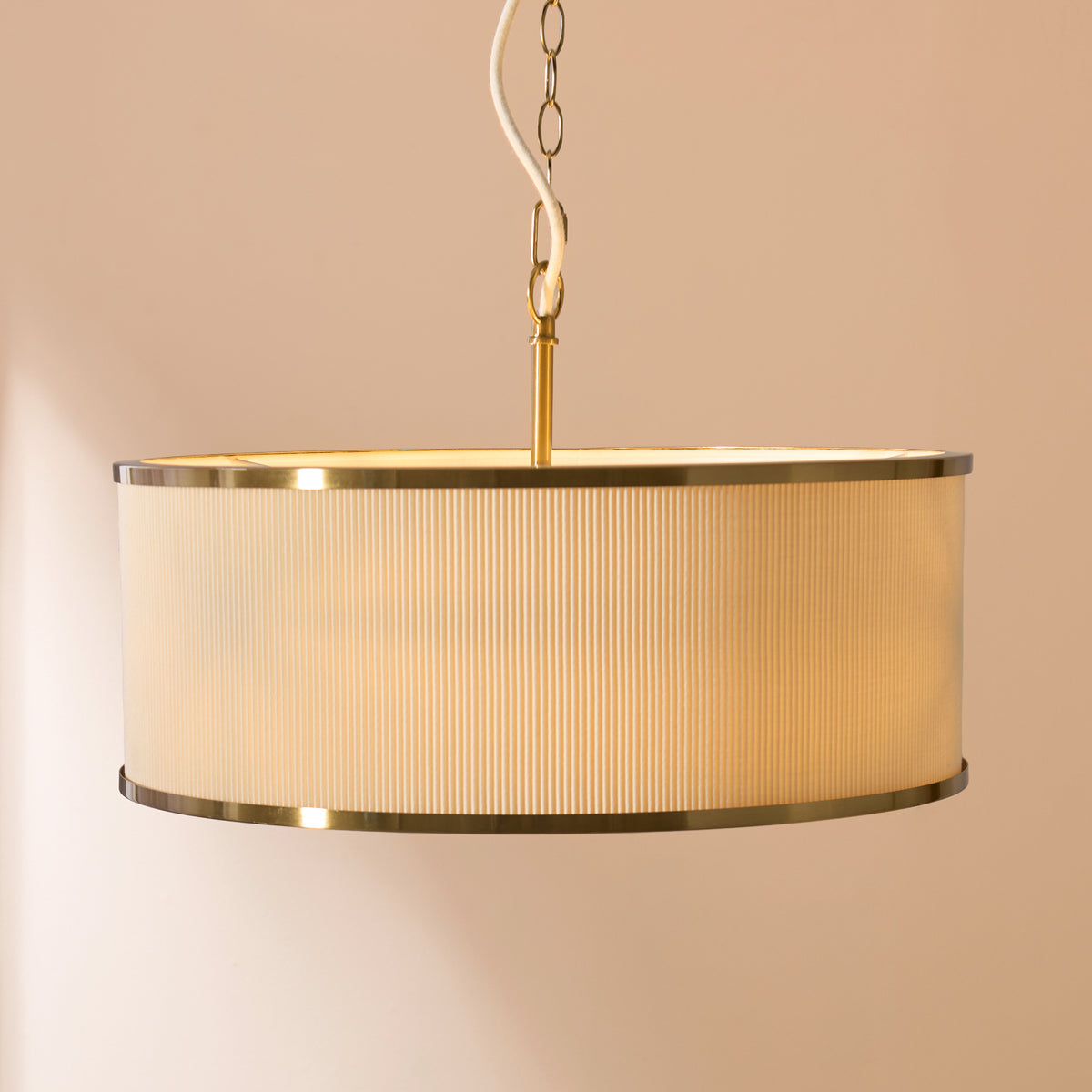 Lexi drum pendant with pleated ivory shade and brass rims glowing against a warm pink wall