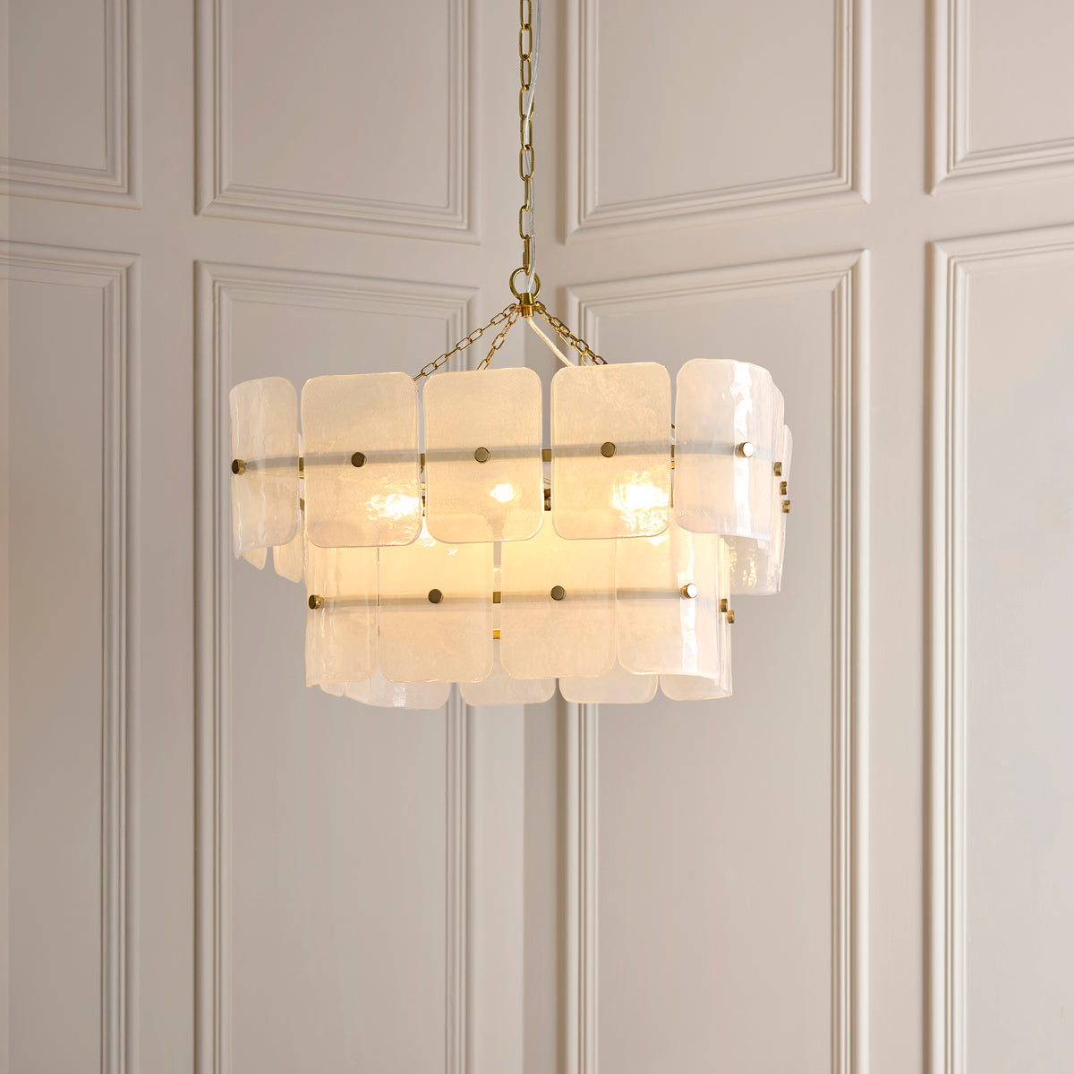 Venice 6 light chandelier glowing against grey panelled walls, its white glass panels brass-studded