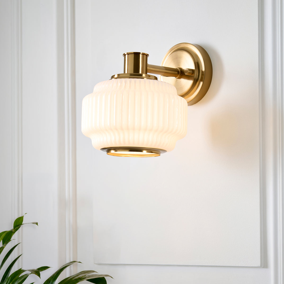 Evora brass wall light mounted on a white wall beside a green plant, opal ribbed shade unlit