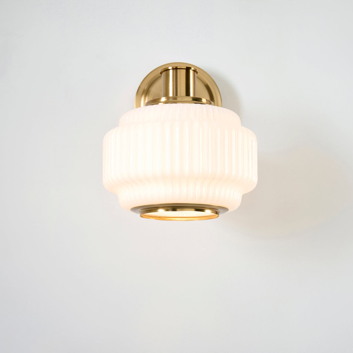 Evora wall light seen head-on with its ribbed opal glass shade and brass trim ring lit