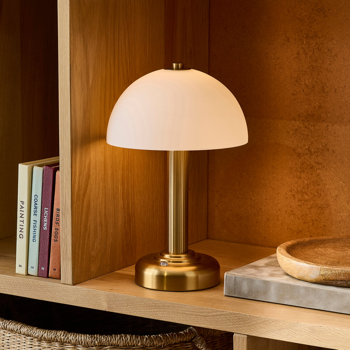 Bridport brass table lamp lit on an oak shelf beside upright books, a wooden bowl and woven basket