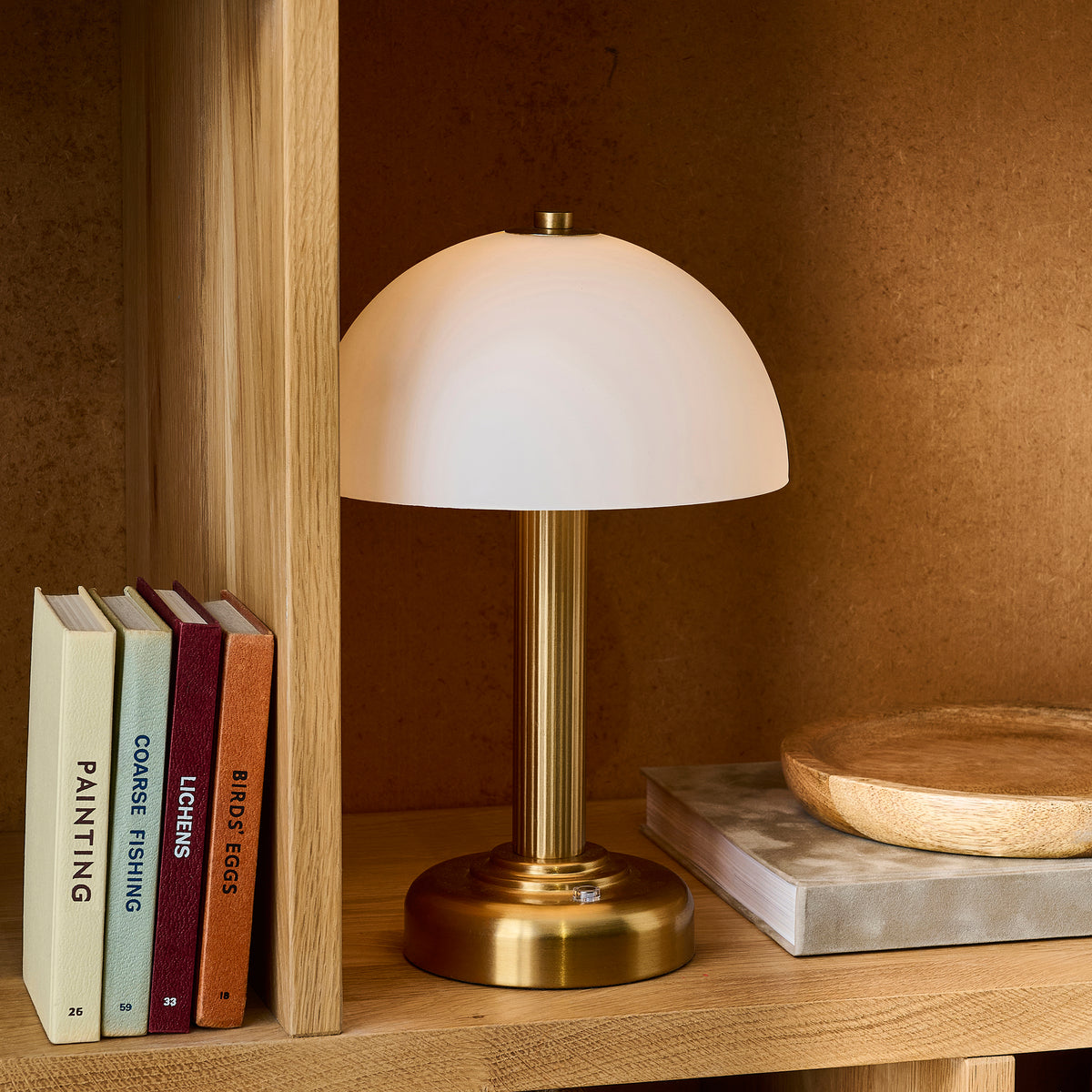 Bridport rechargeable table lamp glowing on an oak bookshelf next to hardback books and a wooden dish