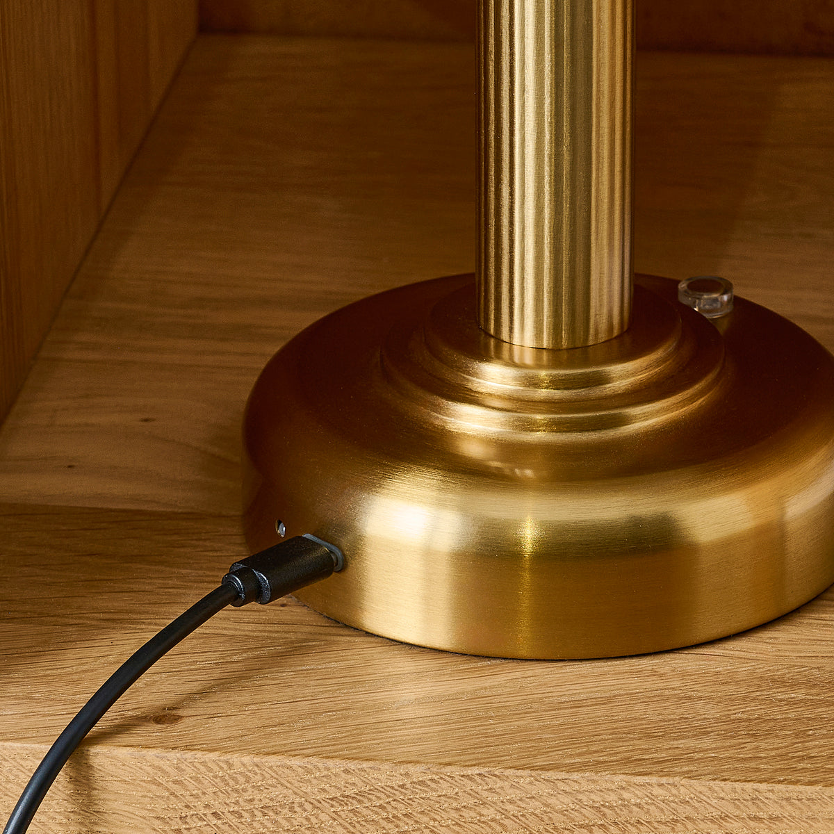 Charging cable plugged into the side of the Bridport lamp's brushed brass stepped base on oak
