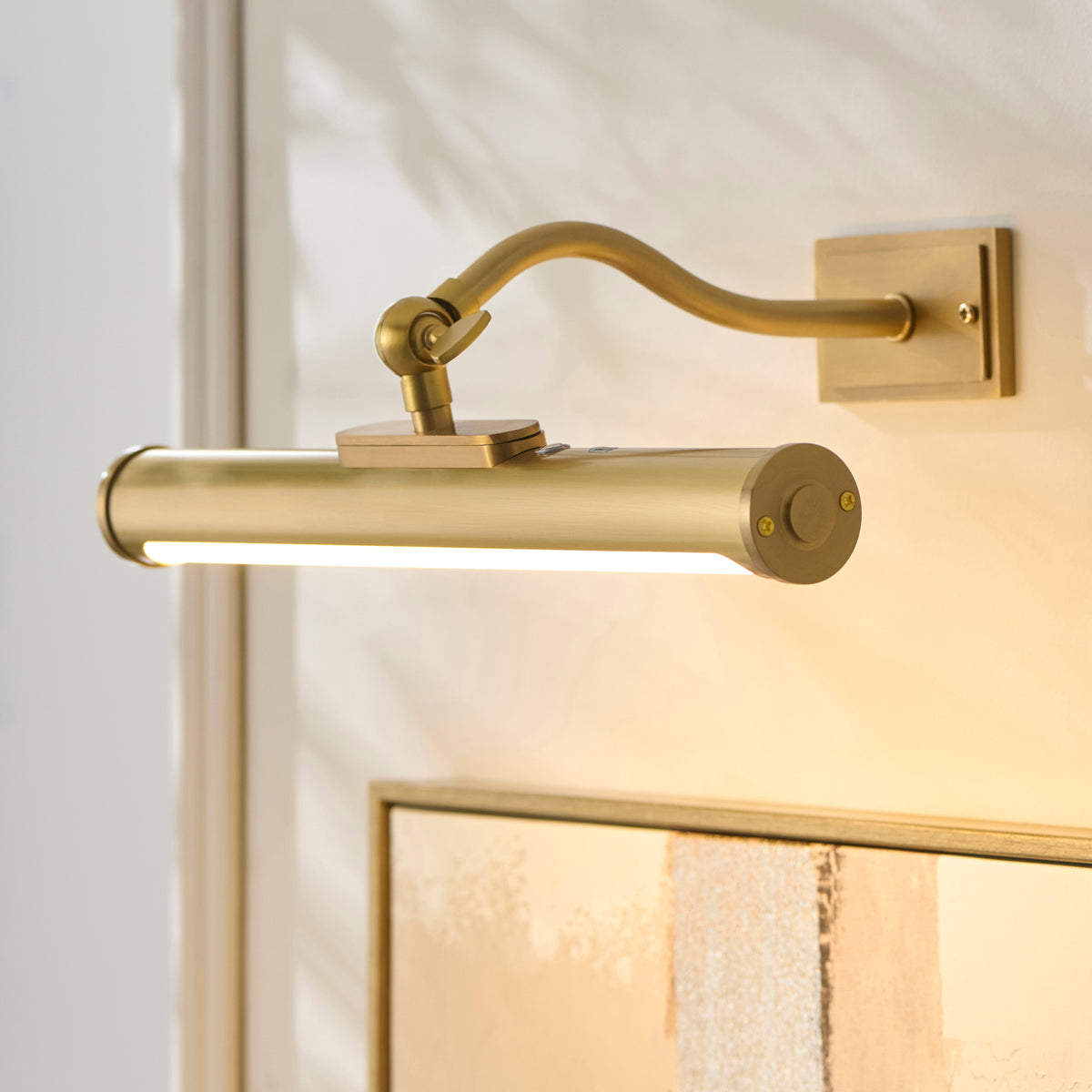 Angled view of the Maris picture light's brass tube and swan-neck arm glowing above a framed artwork