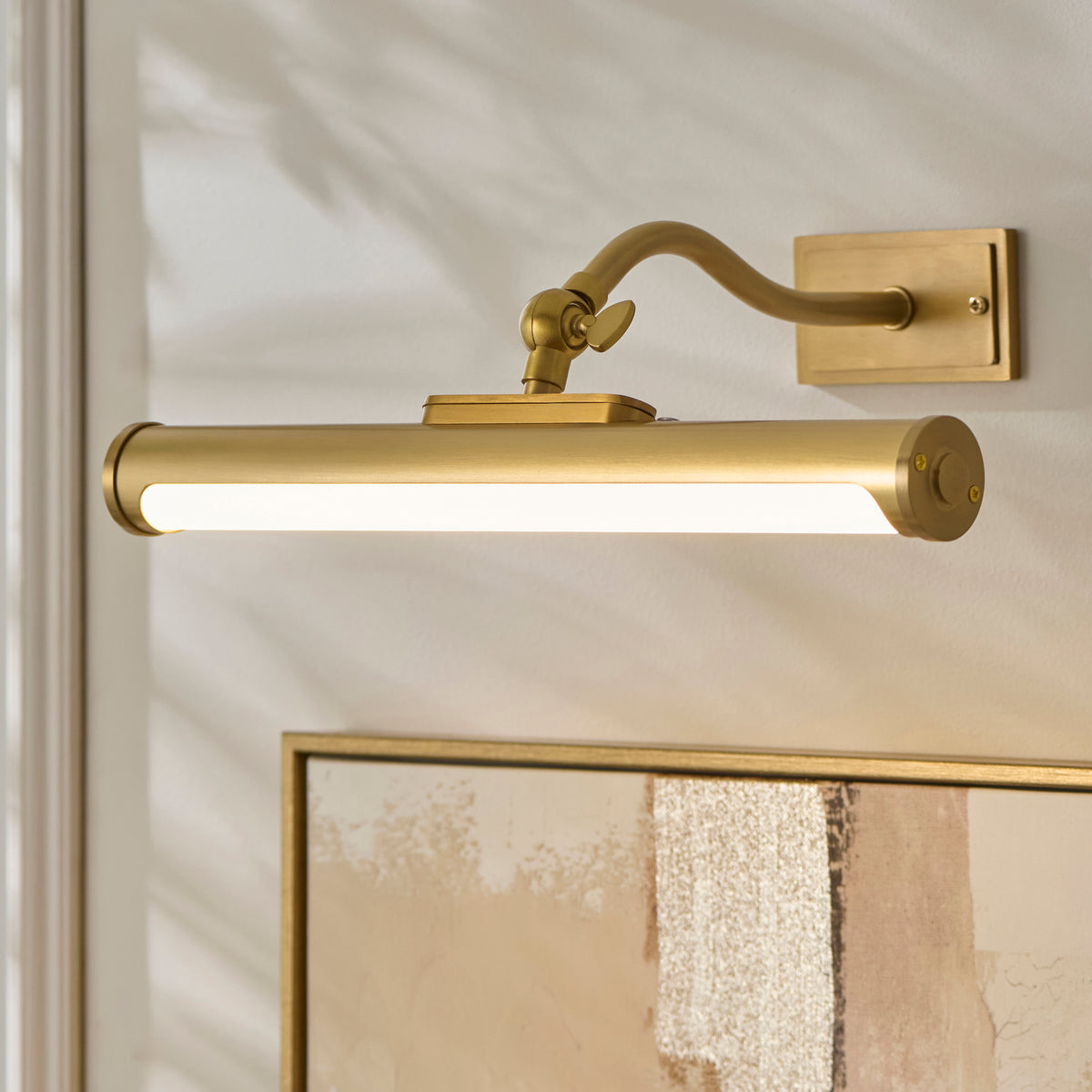 Maris brass picture light angled over a gilt picture frame, its LED strip lit warmly