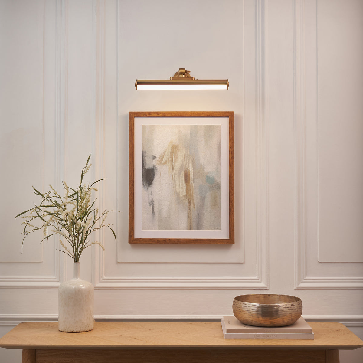 Cirali brass picture light lit above a framed abstract print, console table with vase and brass bowl