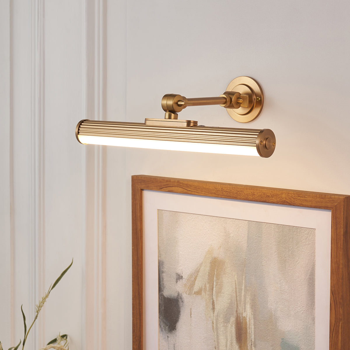 Angled view of the Cirali fluted brass picture light glowing above an oak-framed artwork