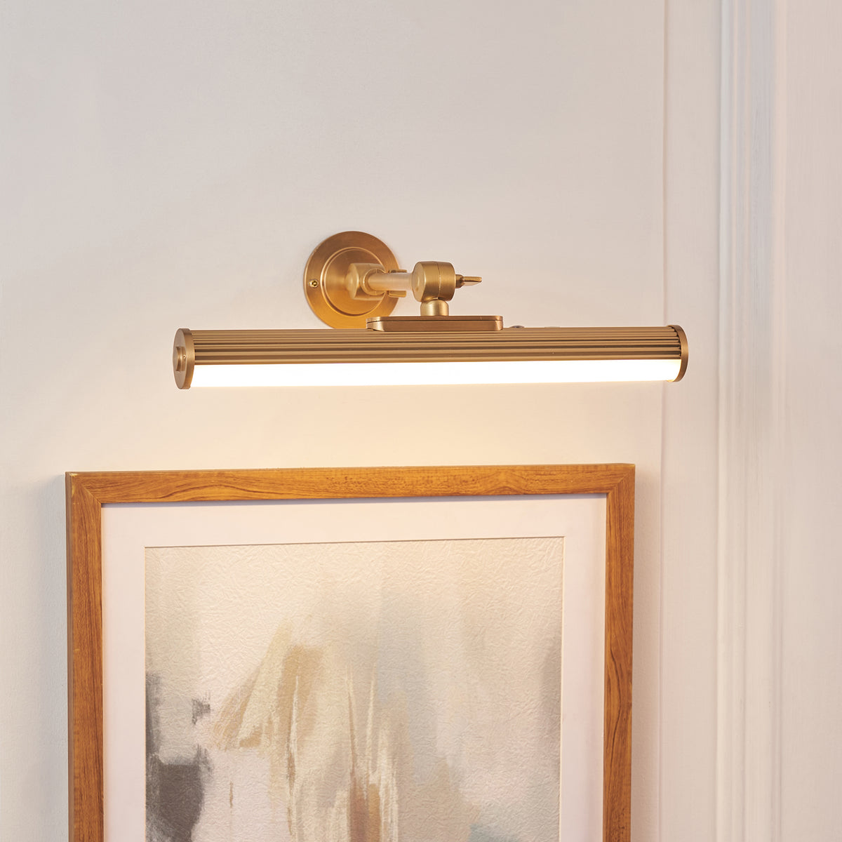 Cirali warm brass picture light with ribbed tube casting light over a wooden-framed print