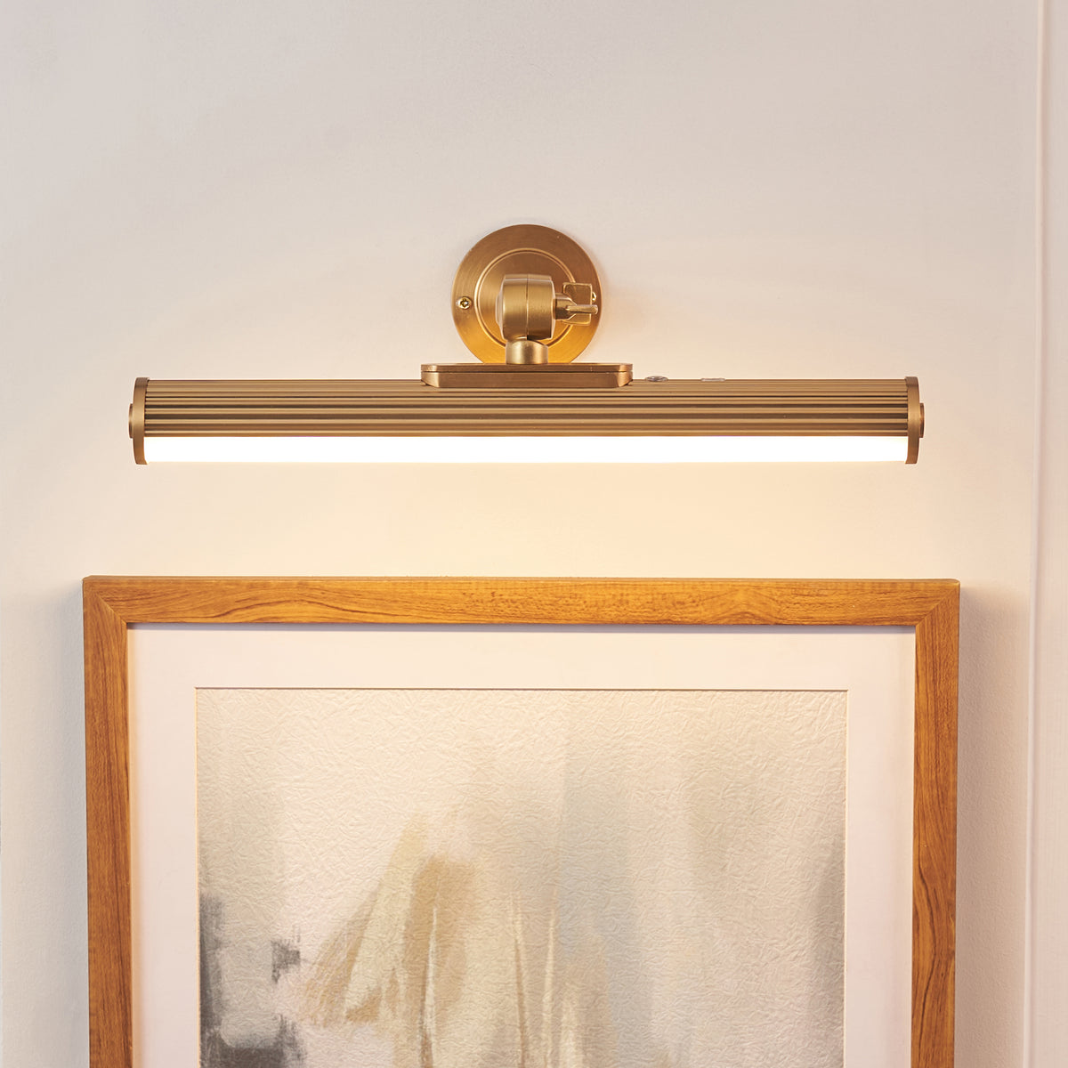 Cirali picture light in warm brass mounted above an oak picture frame, fluted tube softly lit