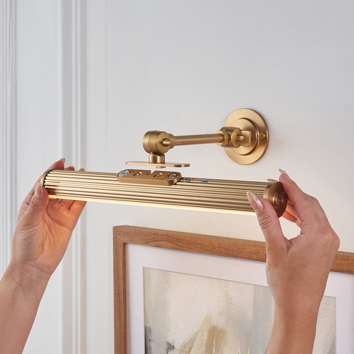 Hands removing the Cirali fluted brass light bar from its wall bracket above a framed print