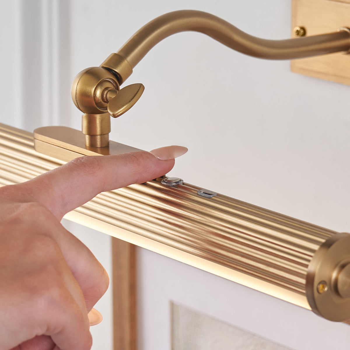 Finger pressing the discreet touch switch on the ribbed brass body of the Kindos picture light