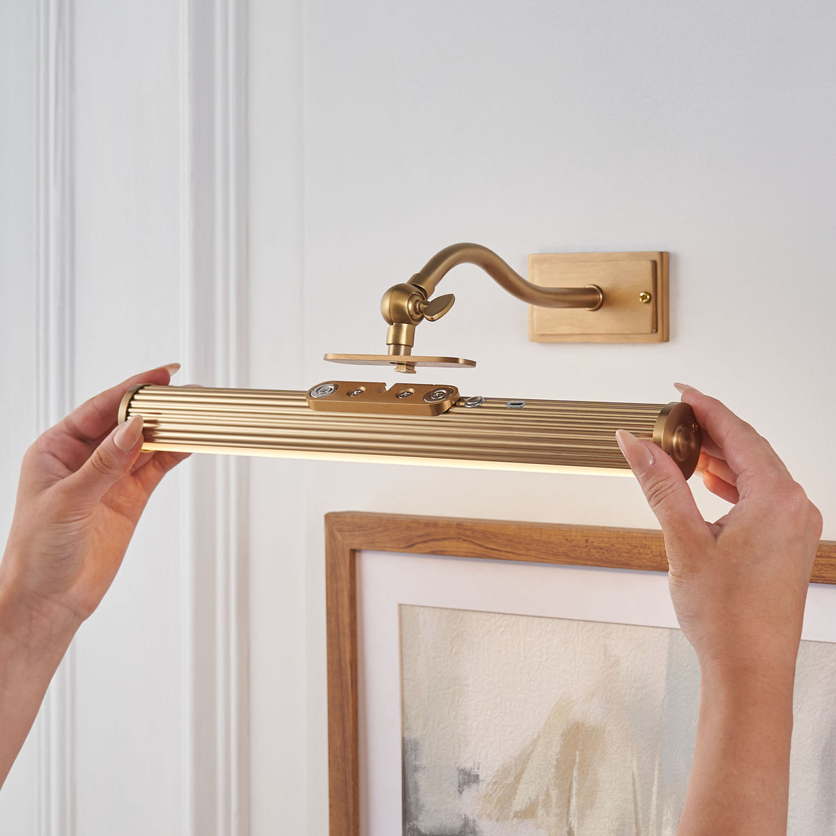 Hands lifting the ribbed brass Kindos light head off its magnetic bracket above a framed print
