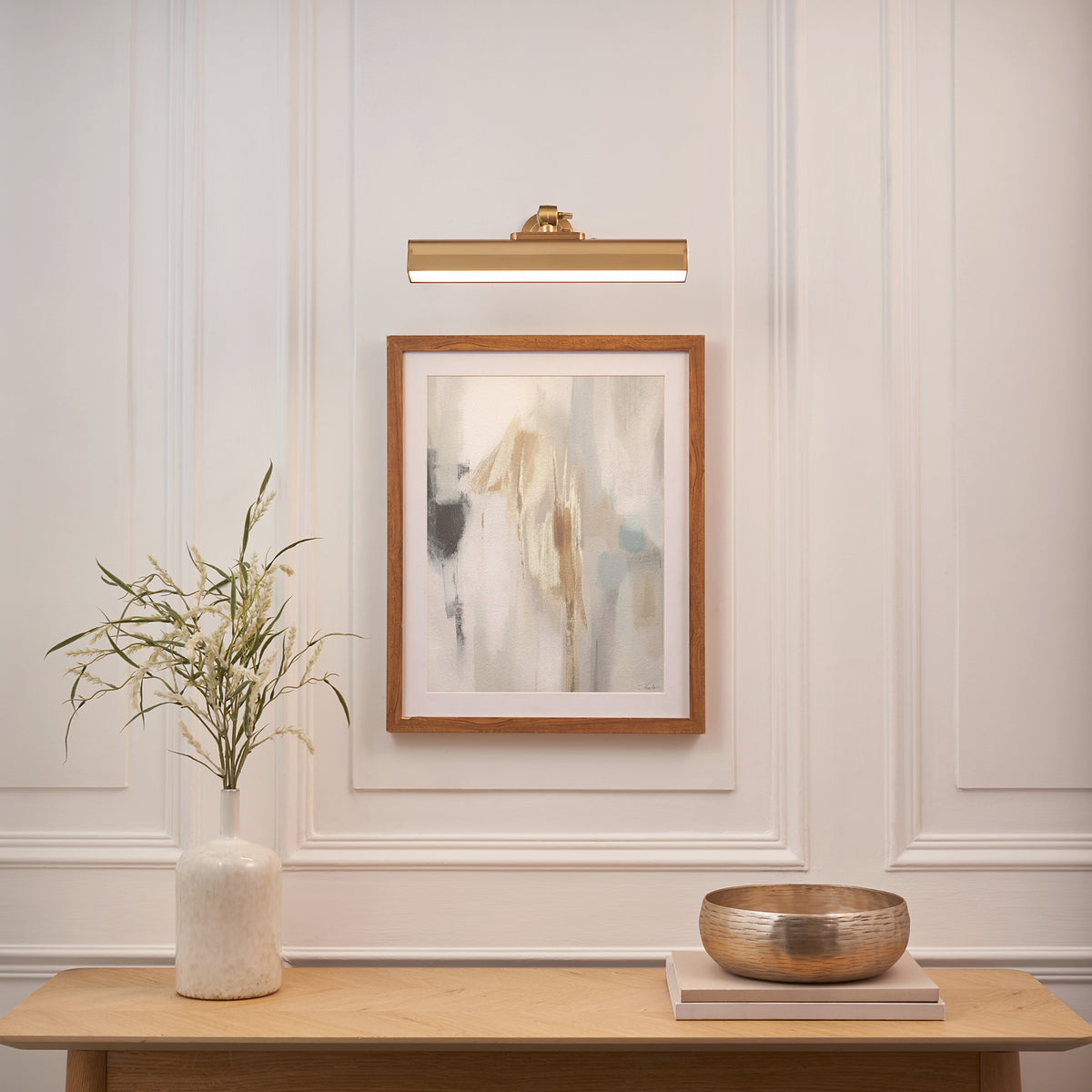Dalyan brass picture light over a framed abstract print, with vase, books and brass bowl below