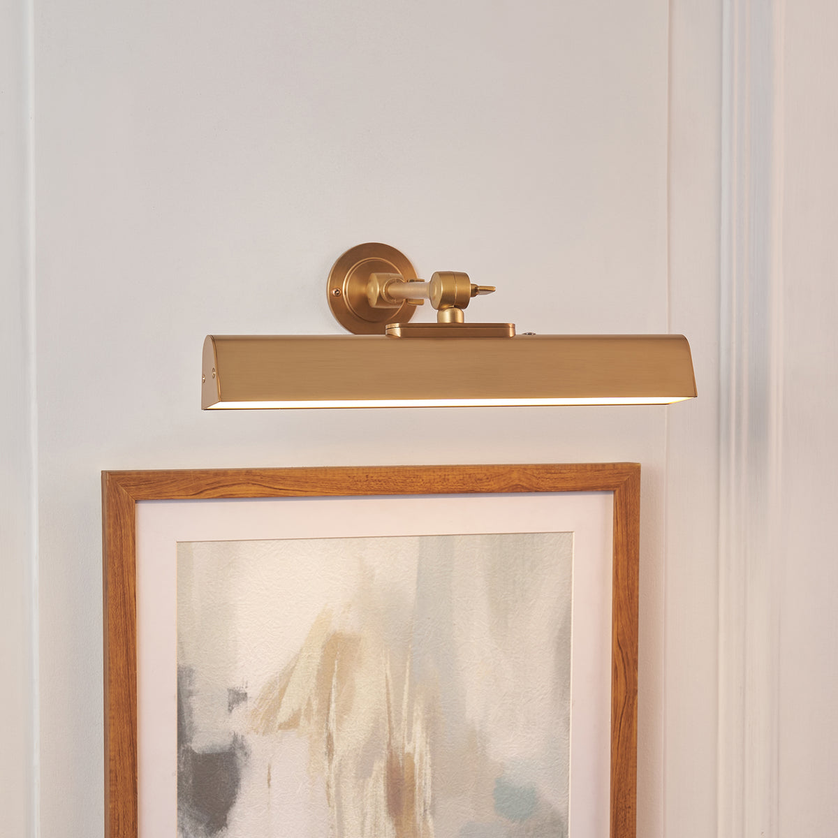 Slim brass Dalyan picture light lit above an oak-framed abstract print on a panelled wall