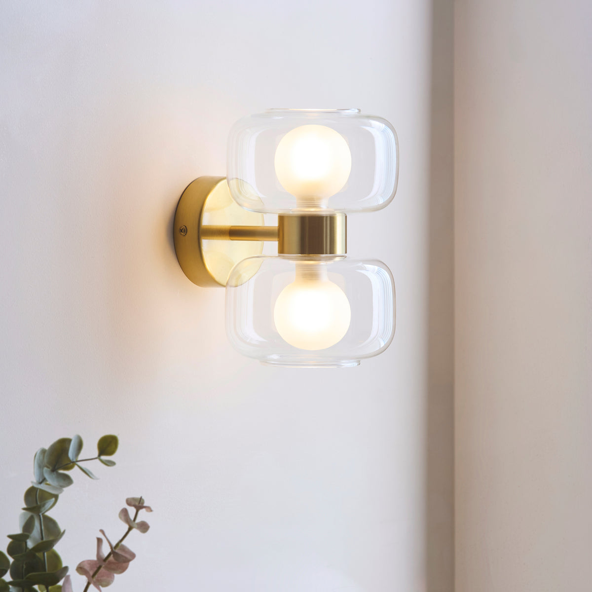 Rossano brass wall light glowing on a pale wall with eucalyptus foliage in the foreground