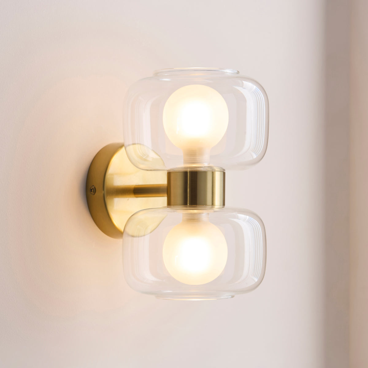 Rossano wall light in brass with twin clear glass shades casting soft light on a white wall