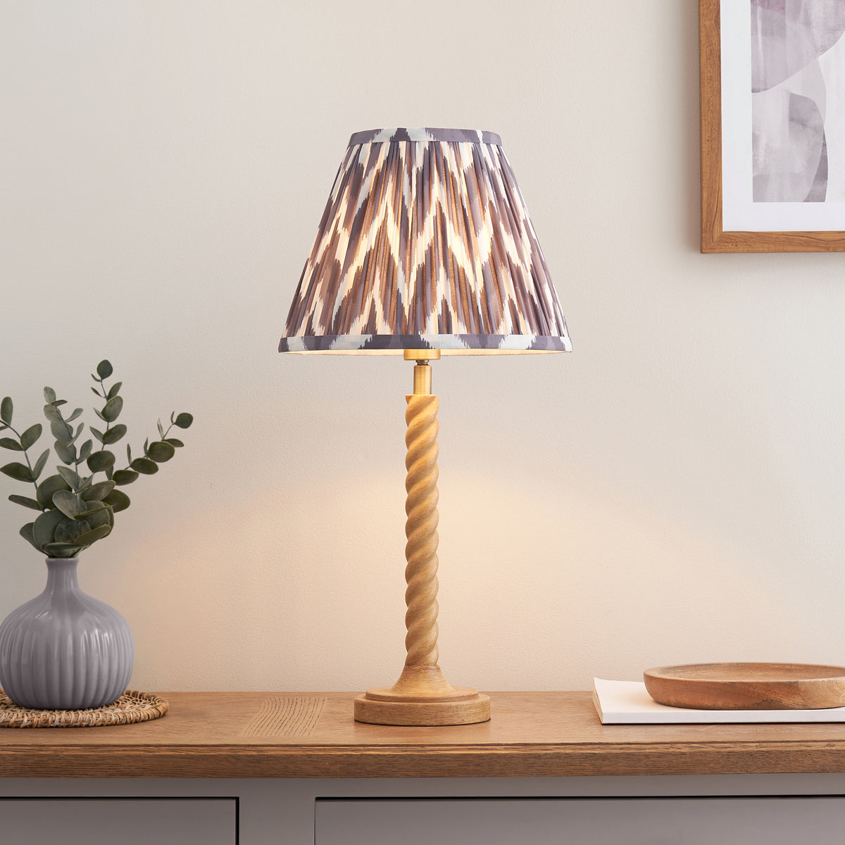 Oak effect twisted table lamp with grey zigzag shade lit on a wooden sideboard beside a vase of eucalyptus