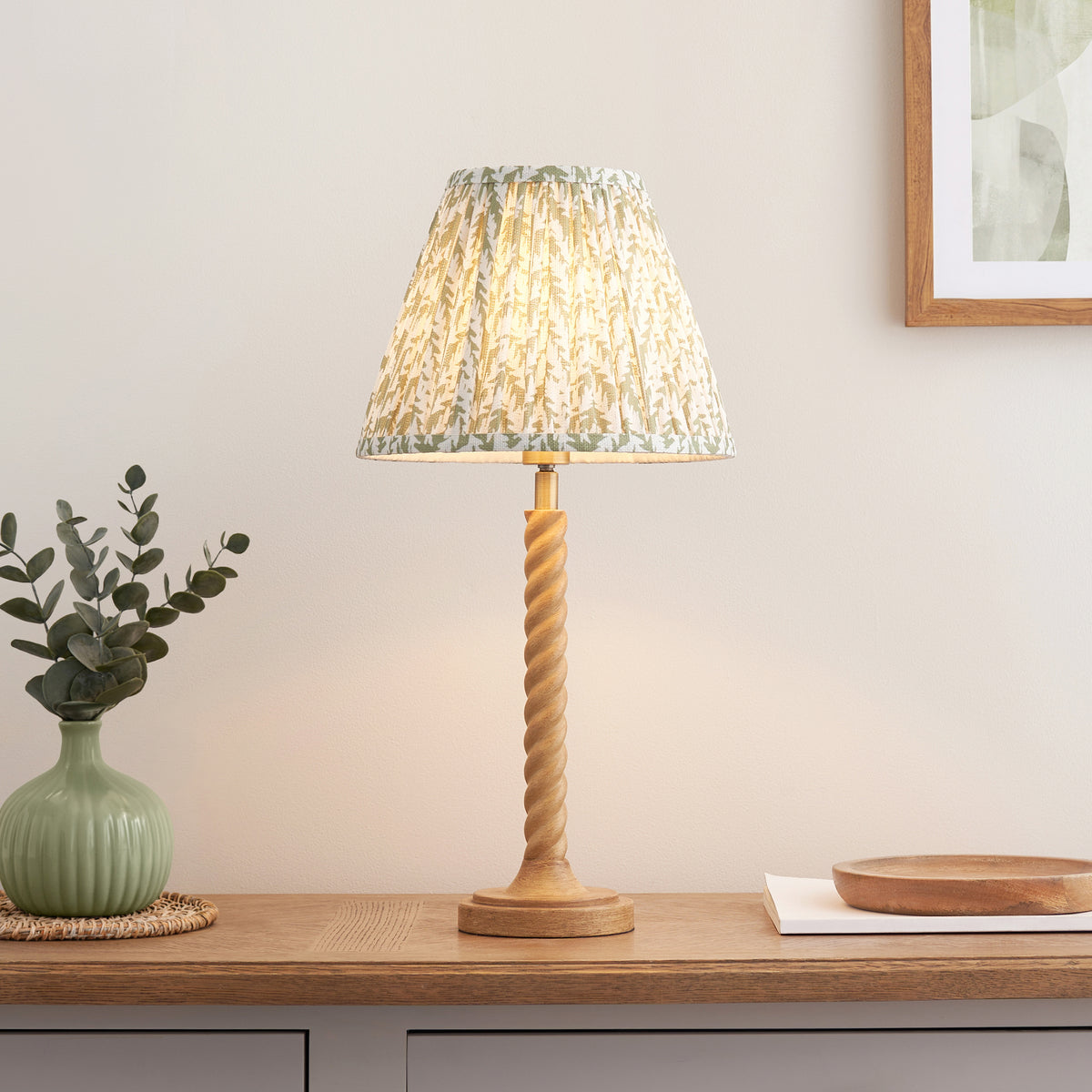 Green leaf print shade table lamp lit on an oak sideboard beside a green vase of eucalyptus