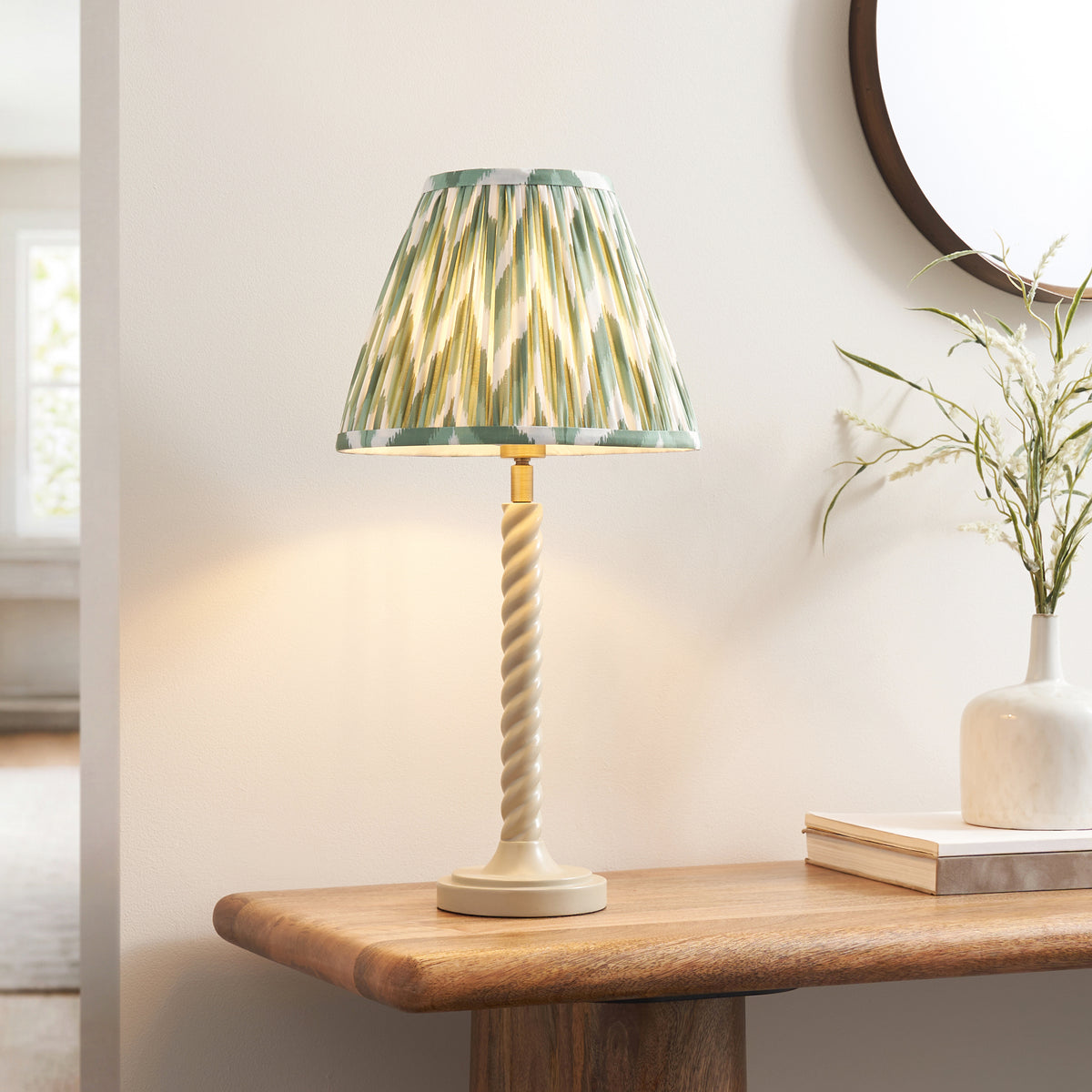 Green zigzag shade table lamp lit on an oak console with books, a white vase and round mirror