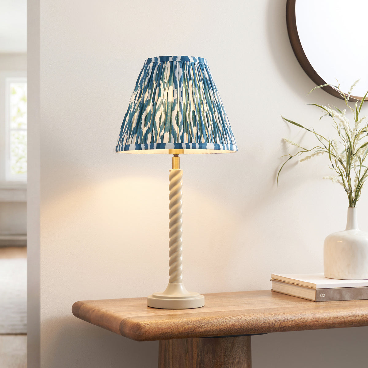 Blue ikat shade table lamp lit on an oak console with books, a white vase and round mirror