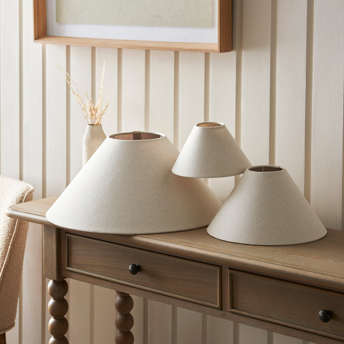 Three natural linen coolie shades in three sizes on a wooden console with dried grasses