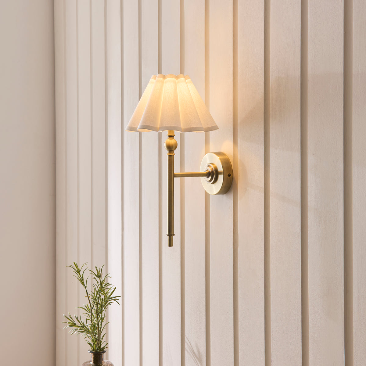 Sylvie antique brass wall light lit on cream panelled walls with a small potted plant below