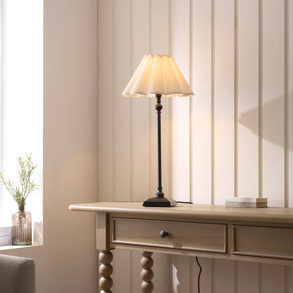 Sylvie black table lamp lit on a pale wooden console table beside a window