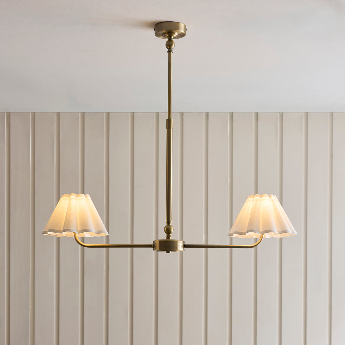 Sylvie antique brass linear pendant with two glowing scalloped shades against a panelled wall