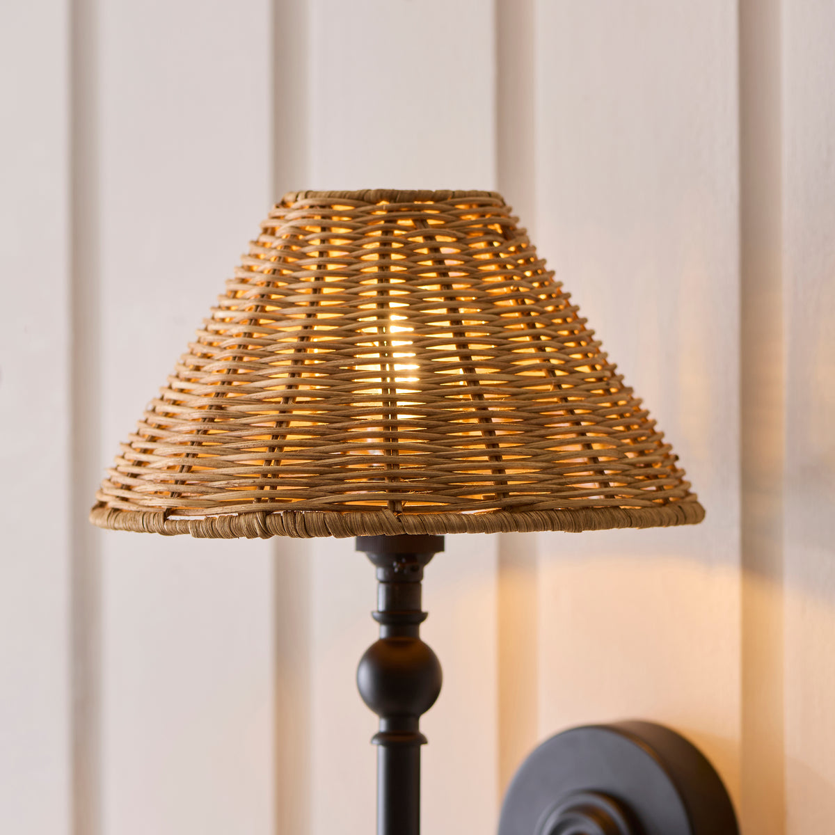 Close-up of the Jericho wall light's woven rattan shade lit above its black turned stem