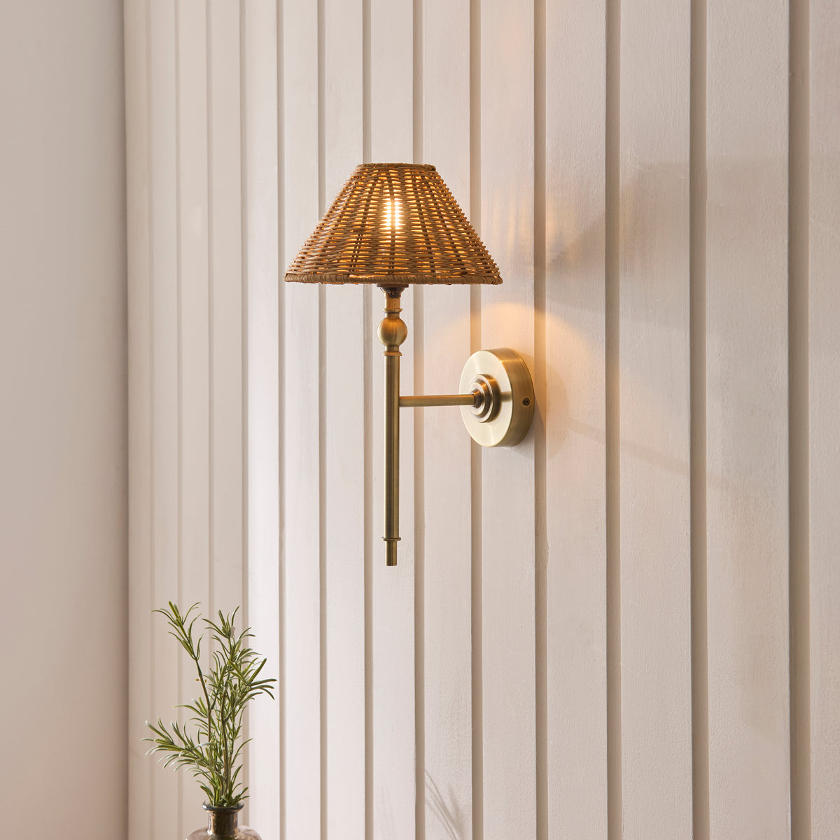 Jericho brass wall light lit on cream panelling beside a small potted rosemary plant