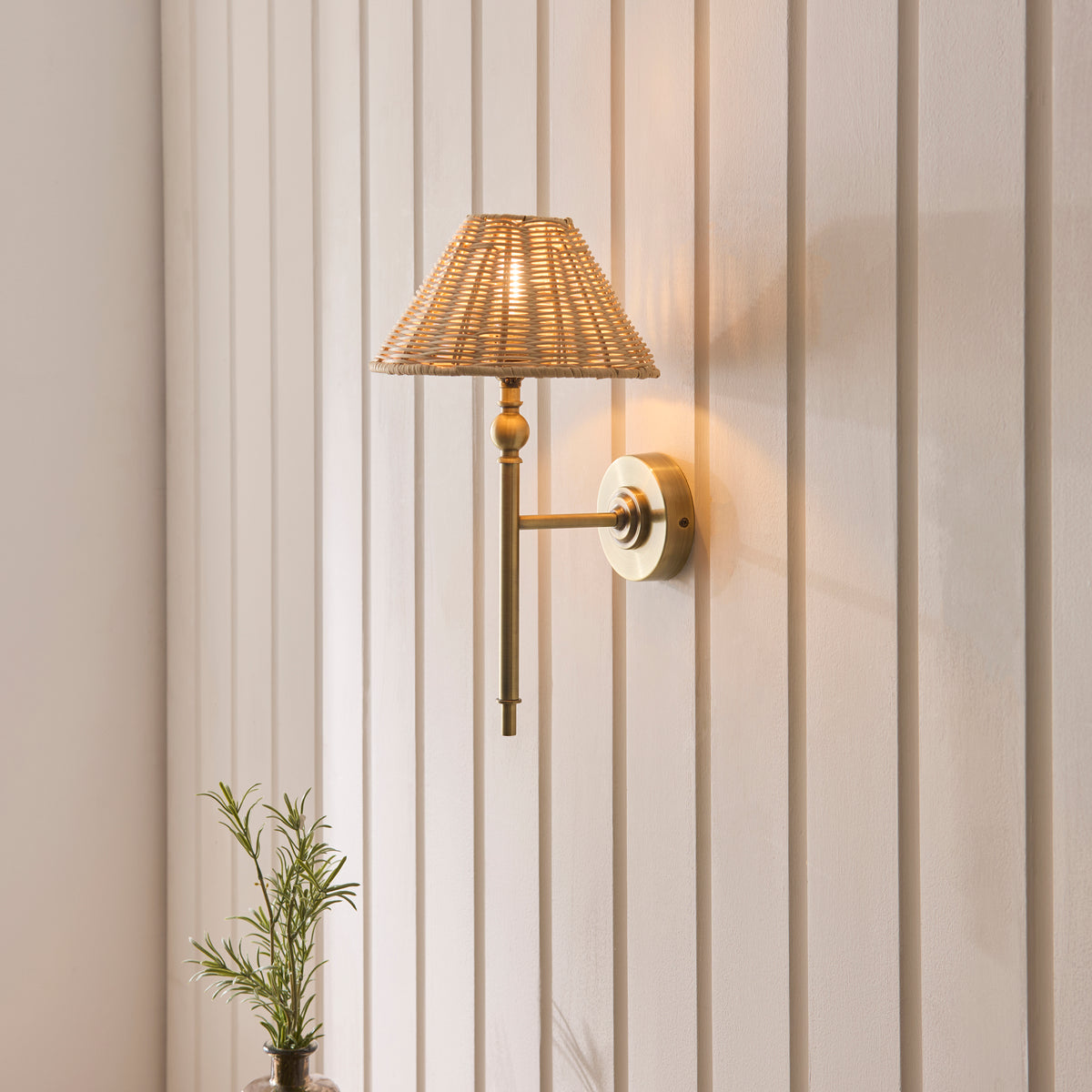 Jericho brass wall light glowing on cream panelling above a potted rosemary plant