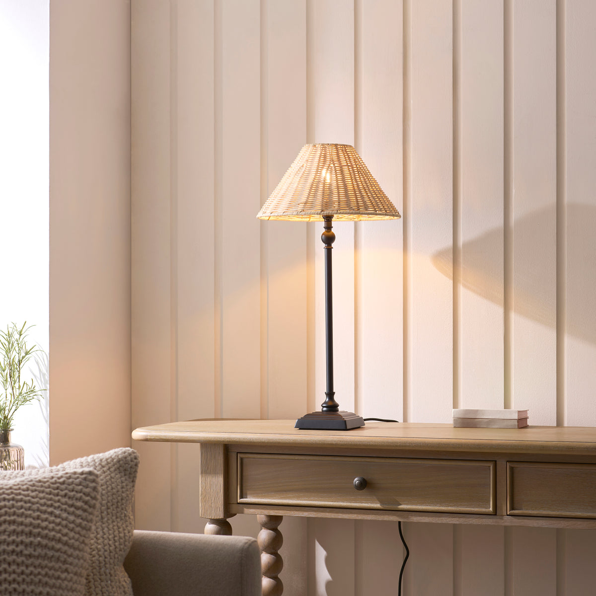 Jericho black rattan table lamp lit on an oak console table beside a cushioned chair