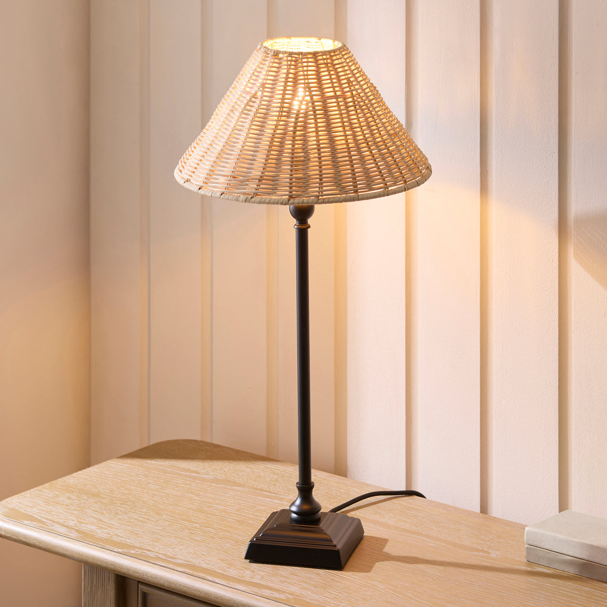 Lit Jericho black table lamp with natural rattan shade on a pale wooden table