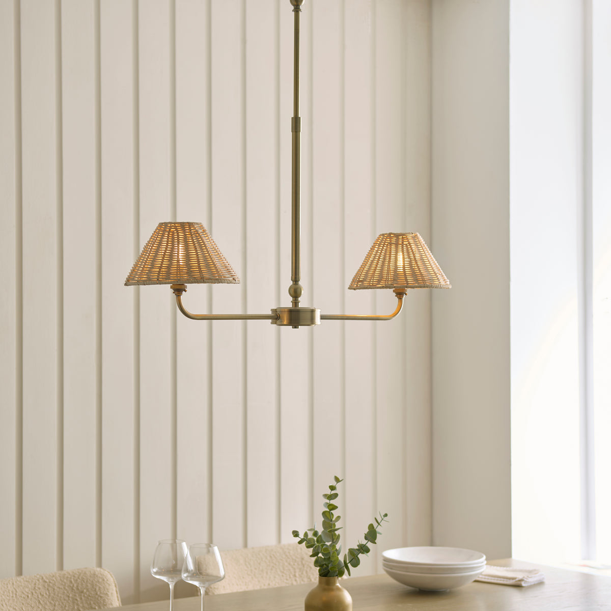 Jericho brass linear pendant lit above a dining table with wine glasses and eucalyptus