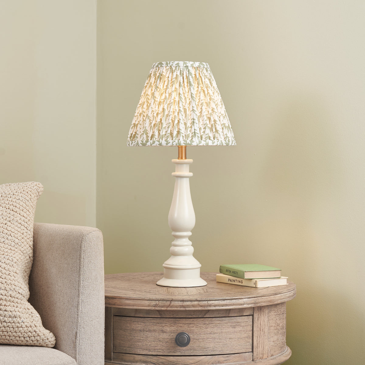 Myrtle white table lamp glowing on a rustic wooden bedside table beside books and a knitted cushion