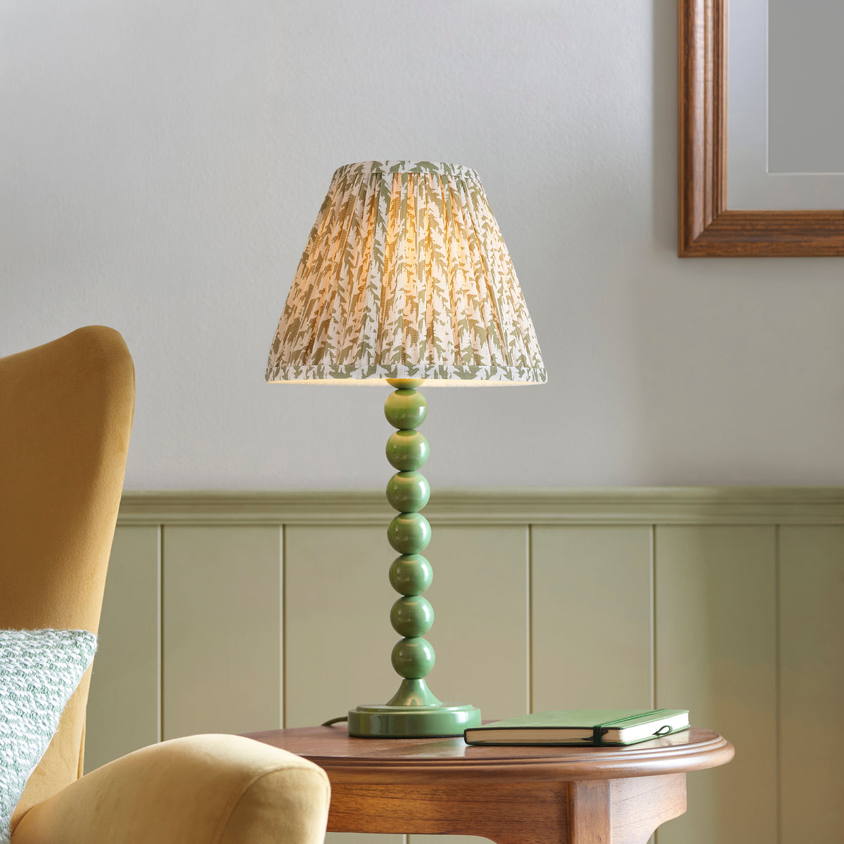 Higgledy green table lamp lit on a wooden side table beside a mustard armchair and book