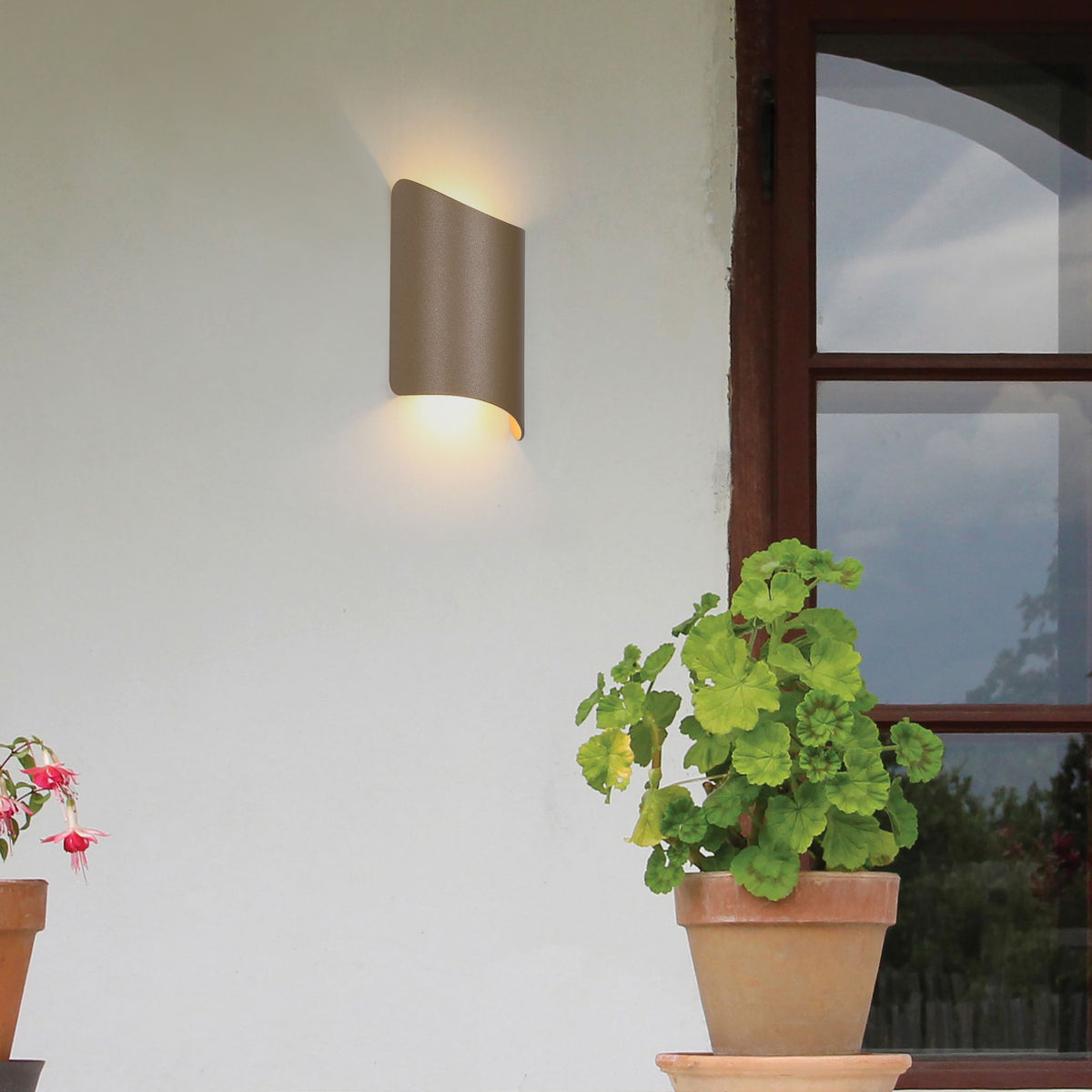 Shard gold wall light lit on a cream house wall beside a window and potted red geraniums