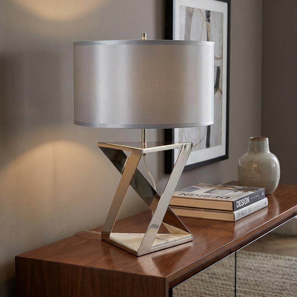 Geometric polished nickel table lamp with a grey drum shade on a wooden sideboard next to books.