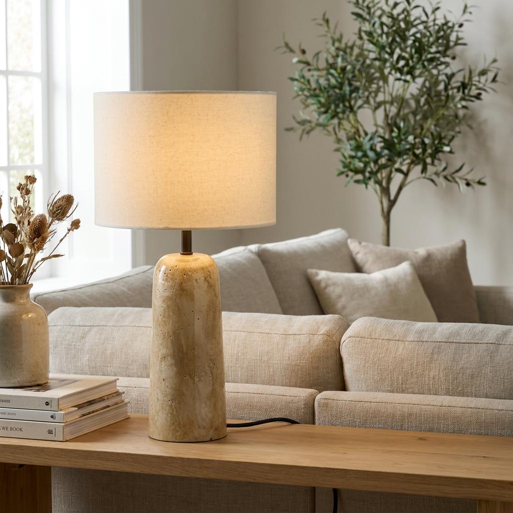 Table lamp with a glazed travertine-effect base and fabric drum shade lit on a wooden console table behind a sofa.