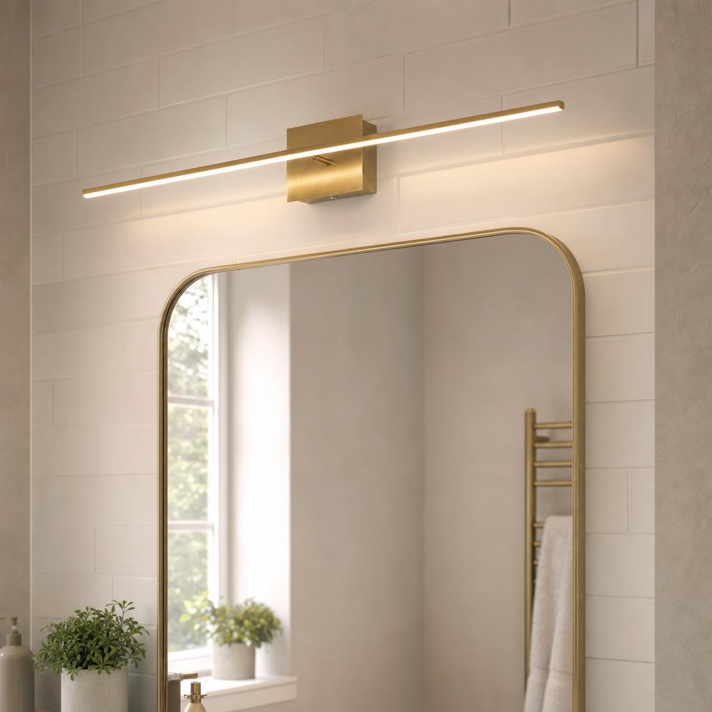 Slim brass LED bar light mounted horizontally above a gold-framed bathroom mirror on a white tiled wall.