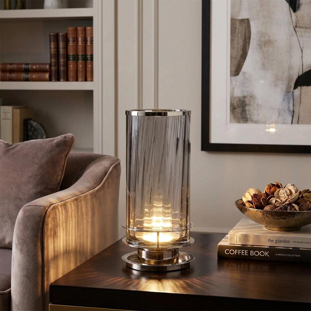 Cylindrical smoked glass table lamp with polished nickel accents glowing on a dark wooden table in a cozy living room.