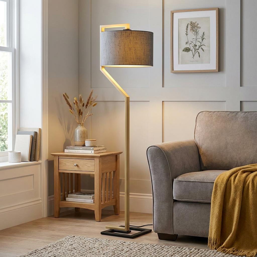 Modern floor lamp with a gold angular base and grey drum shade stands next to a grey armchair in a living room.